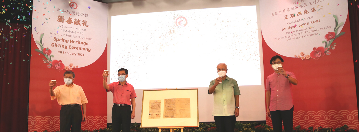 Four masked people toasting, with framed documents in the center and event signage in the background.
